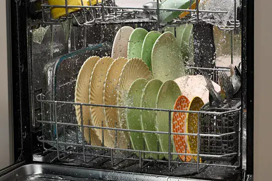 Close-up of a dishwasher lower rack filled with colorful plates being washed, with water droplets visible on dishes and interior walls.