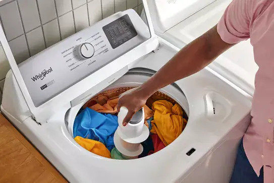 White Whirlpool top-load washer with lid open showing a full load of colorful laundry, with cycle selector dial and controls visible on the panel.