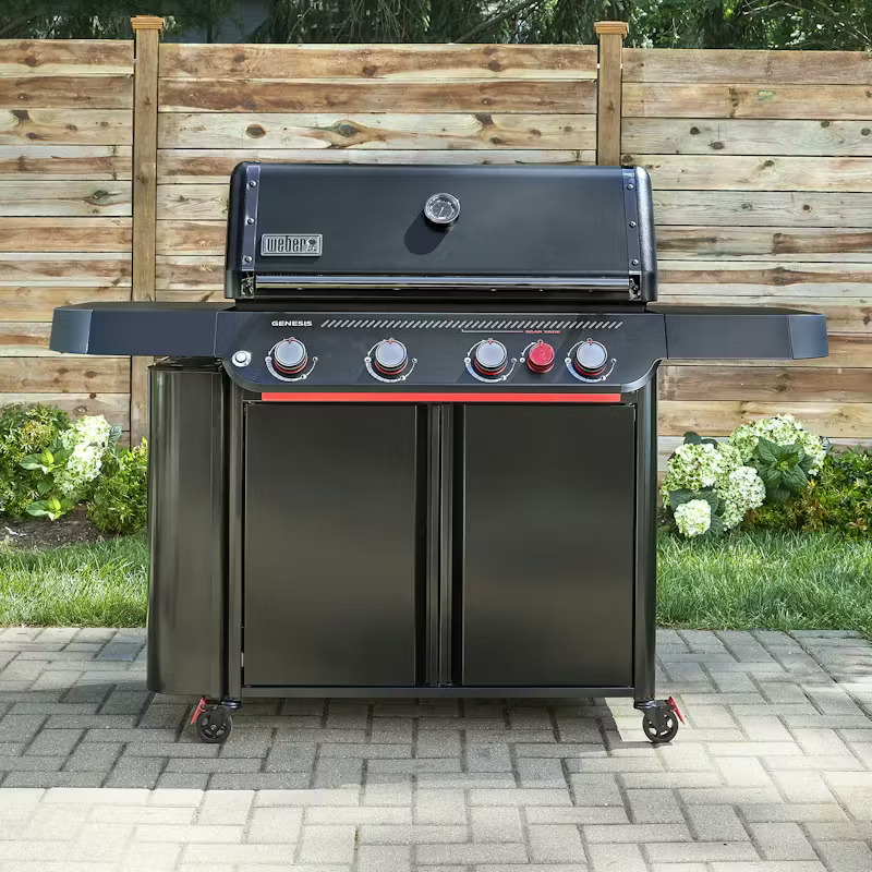 Weber Genesis E-430 Stealth Edition --- with cabinet base and side shelf on a paver patio, with a wood fence and white hydrangeas in the background.