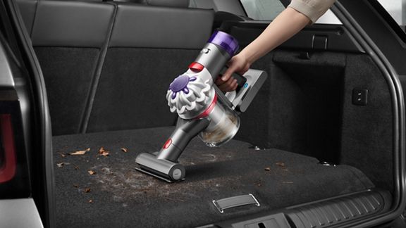 A hand holds a Dyson cordless handheld vacuum cleaning debris and dirt from the carpeted trunk of a car.