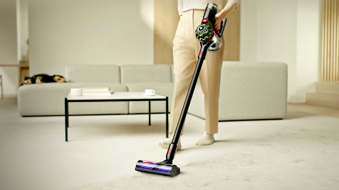 Person in beige trousers using a Dyson V10 cordless stick vacuum cleaner on a light carpet in a modern living room with a grey sofa and coffee table in the background.