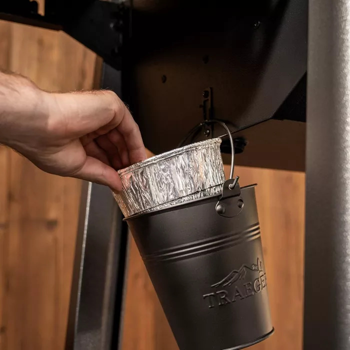A hand removing a foil grease bucket liner from a black Traeger-branded grease bucket mounted to the side of a pellet grill.