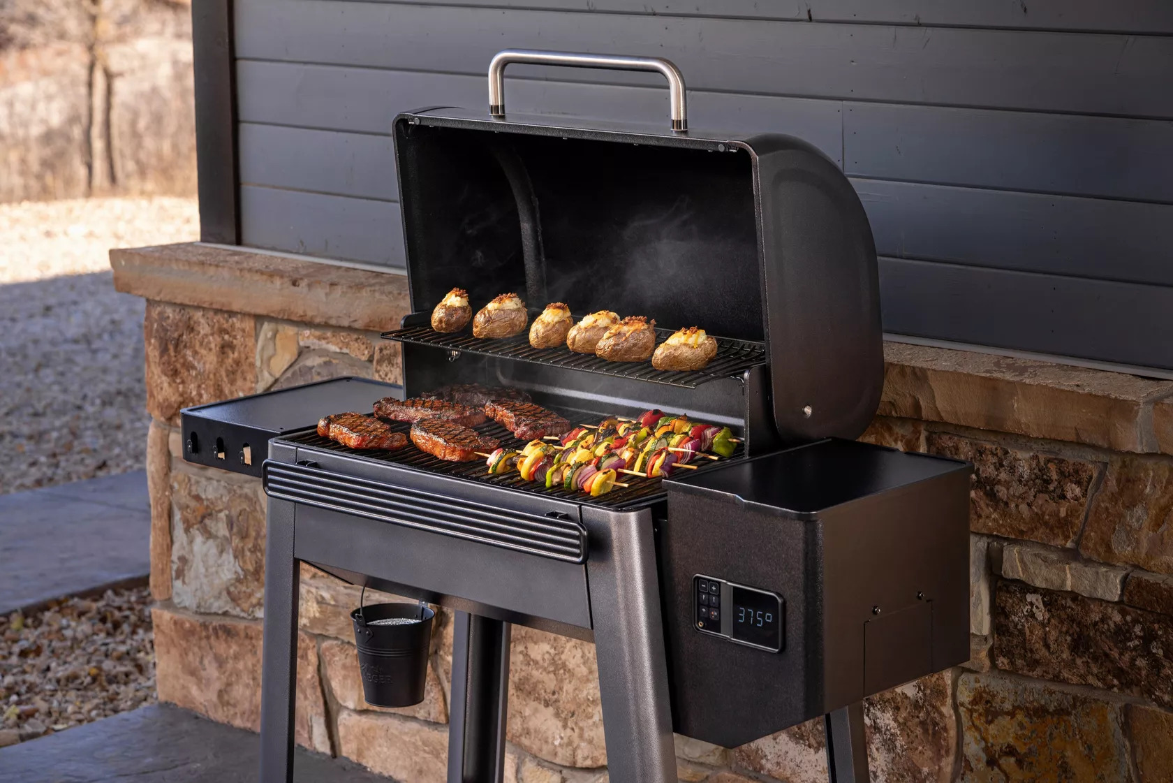 A Traeger Westwood XL pellet grill open at 375 degrees F, cooking steaks, vegetable skewers, and baked potatoes on two grill racks, set on an outdoor stone patio.