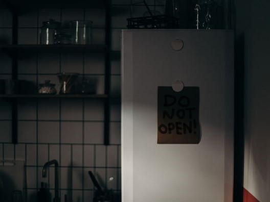 A refrigerator in a darkened kitchen with a note on it that reads DO NOT OPEN